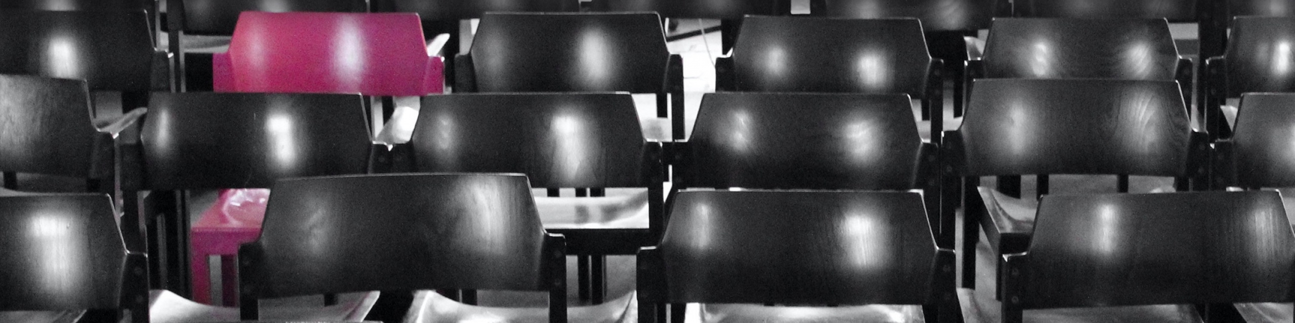 Der pinke Stuhl steht in der Reihe zwischen vielen schwarzen Stühlen.